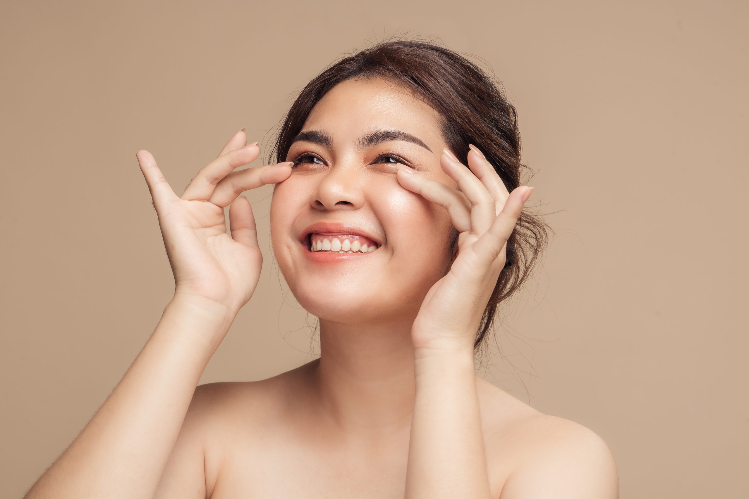 Beautiful smiling Asian female model with natural makeup touching glowing hydrated skin on beige background.